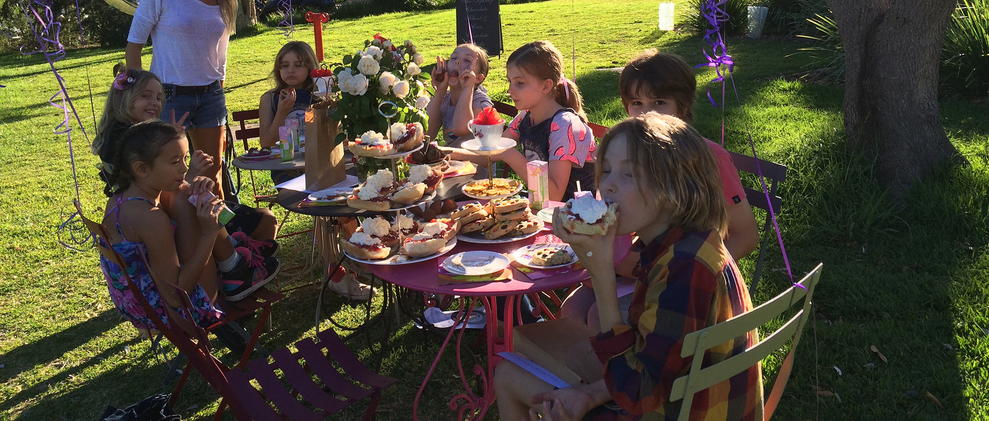 Children at an outdoor birthday table enjoy artisan handmade chocolate cupcakes with white frosting in sunshine
