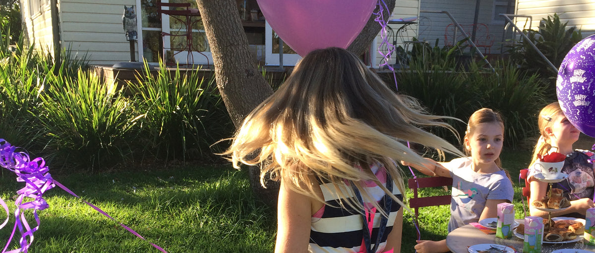 Girl with long hair outdoors at a birthday table, purple balloons and ribbons in the sunny yard