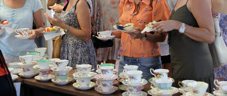People at a tea gathering serve cups from a table lined with patterned teacups and saucers